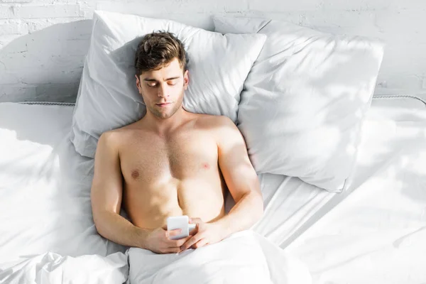 英俊的男人与裸露的躯干躺在床上与智能手机在家里的最高视图 — 图库照片