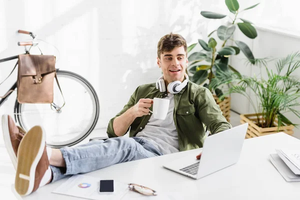 座って オフィスでノート パソコンとテーブルの近くのカップを保持しているヘッドフォンでハンサムな実業家 — ストック写真
