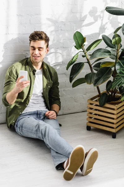 緑のシャツとジーンズのレンガの壁とのオフィスでの植物のそばに座って スマート フォンを保持している男 — ストック写真