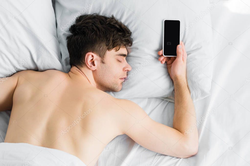 top view of handsome man sleeping under blanket in bad near smartphone with blank screen at home