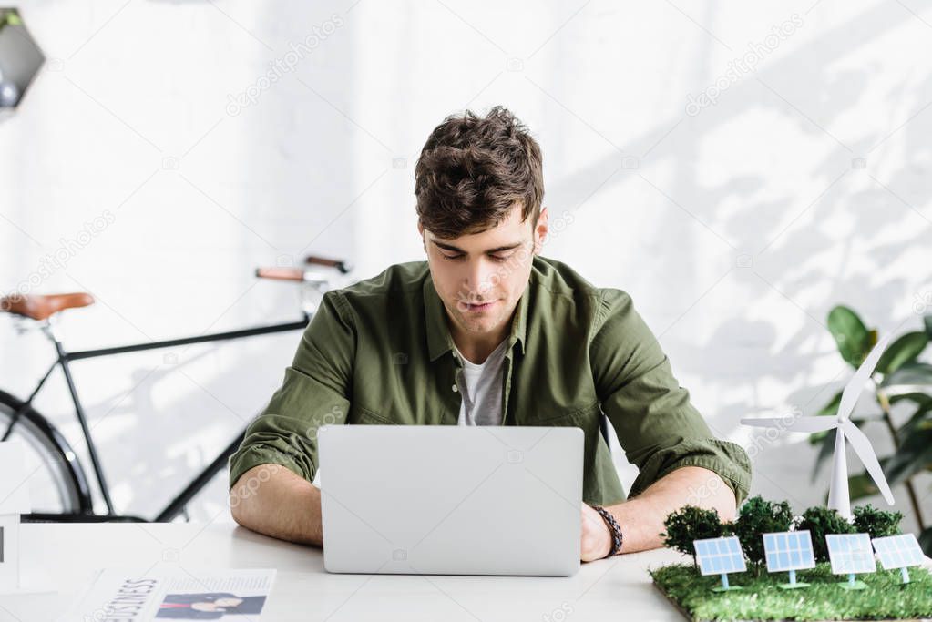 handsome architect in green shirt sitting at table with laptop and windmill, solar panels models in office