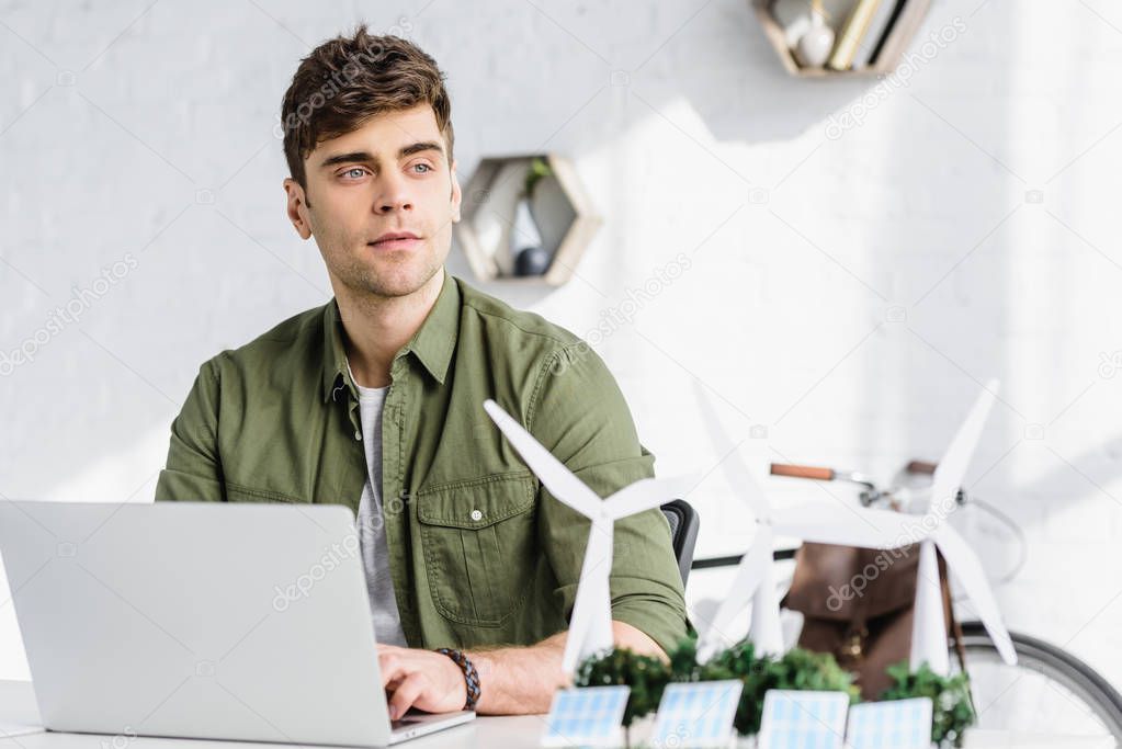 selective focus of handsome architect at table with laptop, solar panels models, windmills and trees models in office