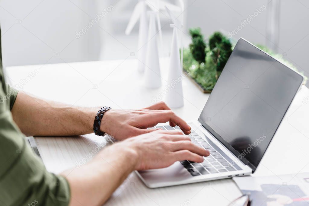 cropped view of architect sitting at table and typing on laptop near windmills models in office