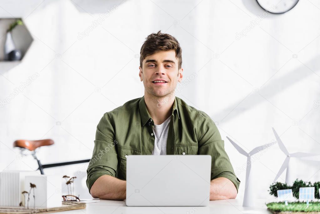 handsome architect sitting at table, typing on laptop and smiling near windmills and buildings models in office