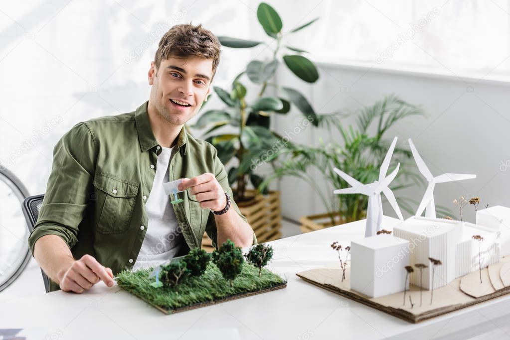 handsome architect smiling and holding solar panels models over grass on table in office