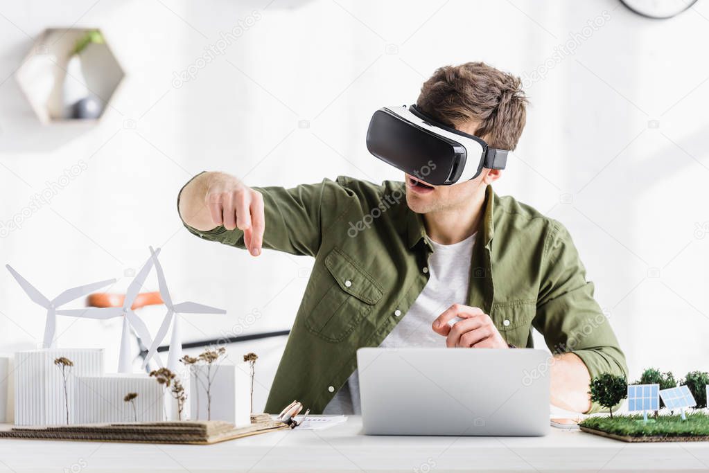 architect in black virtual reality headset sitting at table with laptop and windmills, buildings, trees, solar panels models in office