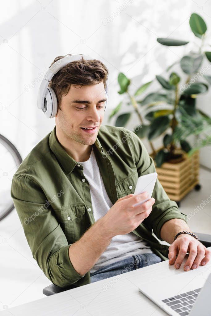 Handsome businessman in shirt and headphones sitting at table with laptop, holding smartphone in office