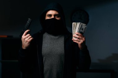 Thief in mask holding gun and dollar banknotes in dark room clipart