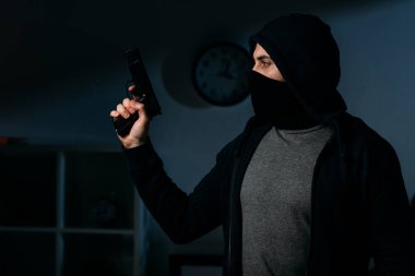Burglar in mask holding pistol in dark room and looking away clipart
