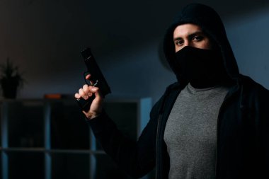 Robber in mask holding gun and looking at camera in dark room clipart