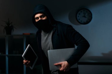 Thief in mask stealing laptop and digital tablet and looking away clipart