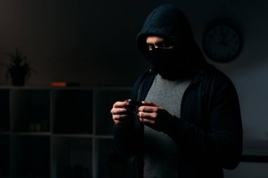 Thief in mask and hoodie standing in dark room and looking at keys clipart