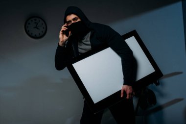 Thief in mask talking on smartphone while stealing flat-screen tv with blank screen clipart