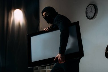 Robber in black mask stealing flat-screen tv with blank screen from apartment clipart