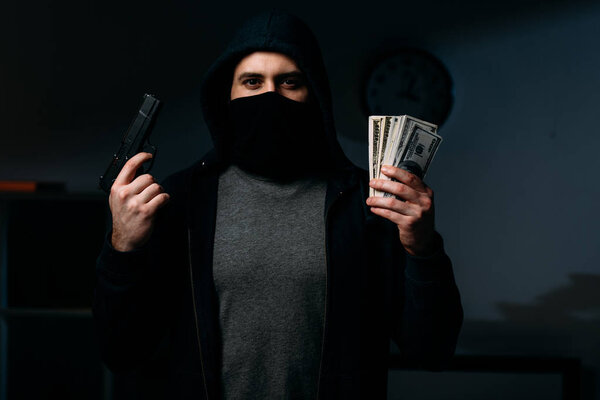 Thief in mask holding gun and dollar banknotes in dark room