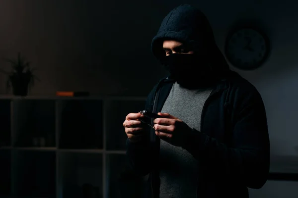 戴面具和头套的小偷站在黑暗的房间里看着钥匙 — 图库照片