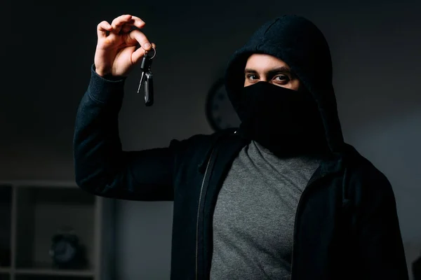 Robber in mask and hoodie holding keys and looking at camera