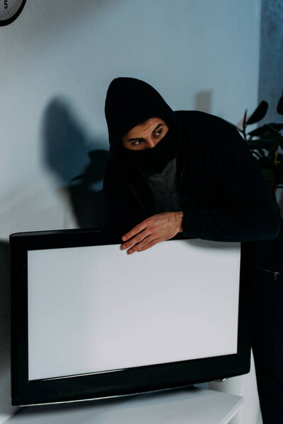 Suspicious thief in mask stealing flat-screen tv with blank screen