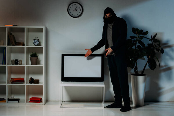 Thief in mask and hoodie pointing with hands at flat-screen tv with blank screen