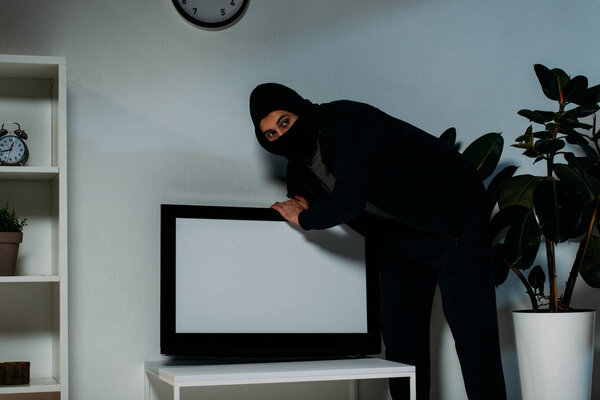 Suspicious robber in mask stealing flat-screen tv with blank screen