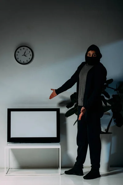 穿着黑色面具的惊讶小偷用双手指着平板电视和空白屏幕 — 图库照片