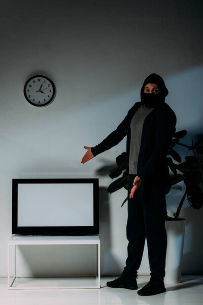 Surprised thief in black mask pointing with hands at flat-screen tv with blank screen