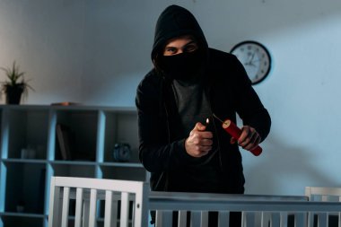 Criminal in mask igniting dynamite near crib and looking at camera clipart