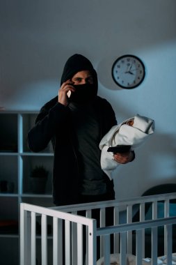 Kidnapper in mask holding infant child and talking on smartphone clipart