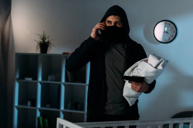 Kidnapper in mask with gun holding child and talking on smartphone clipart