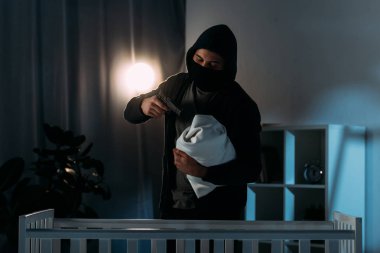 Kidnapper in mask aiming gun at infant child in dark room clipart