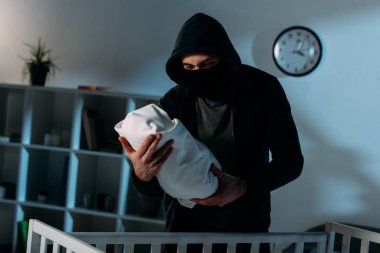 Maske ve siyah hoodie ayakta beşik ve holding bebek çocuk kaçıran