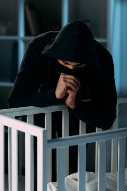 Kidnapper in black hoodie looking in crib and showing please gesture clipart