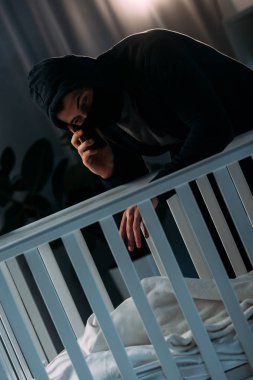 Kidnapper in mask talking on smartphone while standing near crib clipart