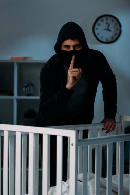 Kidnapper in mask standing near crib and showing hush sign clipart