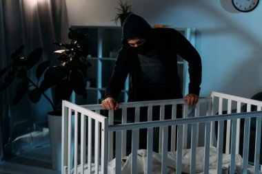 Kidnapper in mask and hoodie looking in crib in dark room clipart