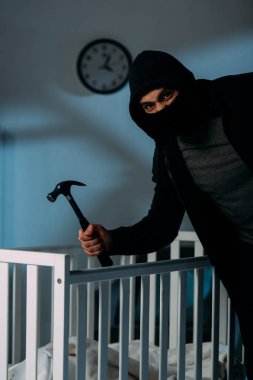 Angry criminal in mask standing near crib and holding hammer clipart
