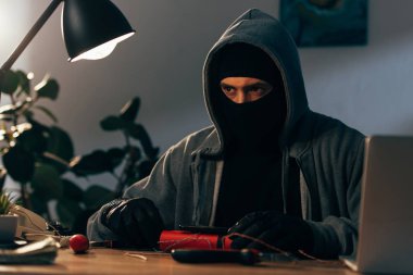 Pensive terrorist in mask and gloves making bomb in room clipart