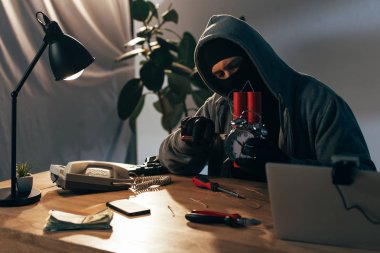 Criminal in mask sitting at table with laptop and making bomb clipart