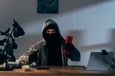 Angry terrorist in mask holding gun and dynamite while sitting at desk in dark room clipart