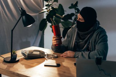 Terrorist in mask sitting in room and holding dynamite clipart