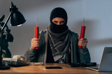 Front view of terrorist with dynamite sitting at table in room clipart