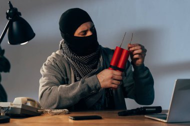 Terrorist in mask holding dynamite while sitting at table in dark room clipart