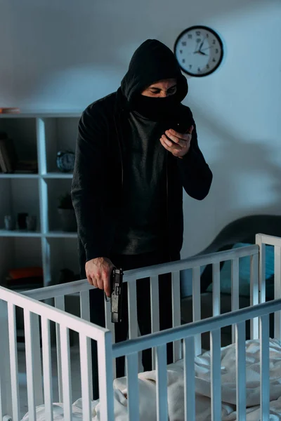 戴面具的罪犯在智能手机上说话 在婴儿床里瞄准枪 — 图库照片