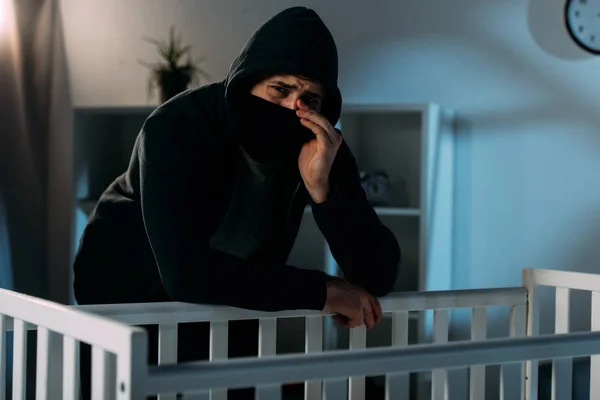 在黑暗的房间里 戴着面具的哭闹绑匪站在婴儿床附近 — 图库照片