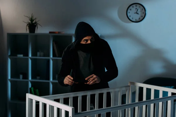 戴着黑色面具的担心的绑匪站在婴儿床附近拍照 — 图库照片