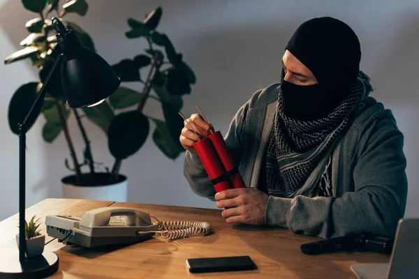 戴面具的恐怖分子坐在桌旁 房间里拿着炸药 — 图库照片