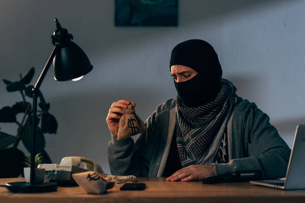 Upset terrorist in mask sitting in room and holding small money bag