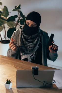 Terrorist in black mask showing gun in video chat in room clipart