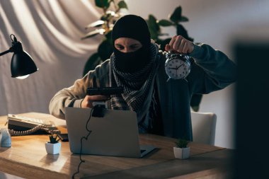 Aggressive terrorist in mask holding gun and showing alarm clock at webcam clipart