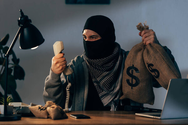 Агрессивный террорист в маске держит мешки с деньгами и смотрит на телефон
